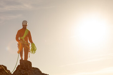 A man in an orange jacket is standing on a rock with a yellow rope. He is wearing a helmet and he...