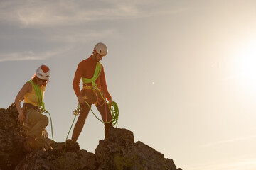 Two people are climbing a mountain, one of them wearing a red jacket. The sun is shining brightly,...
