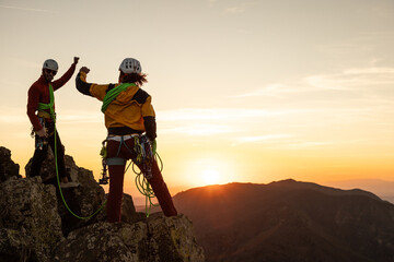 Two people are standing on a mountain top, one of them is wearing a yellow jacket. They are both wearing helmets and are high up on a rock. The sun is setting in the background, creating a beautiful