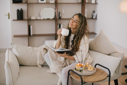 Young woman reading book and drinking tea, sitting on sofa in a cozy living room.