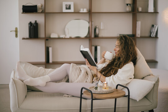 Young woman reading book and drinking tea, sitting on sofa in a cozy living room.