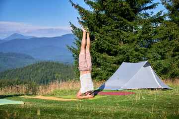 Man performing headstand, yoga pose Sirsasana on a yoga mat in a scenic outdoor setting. Person in grassy area, gray camping tent on background, under clear blue sky.
