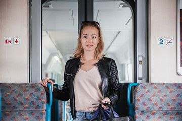 Young blond woman in jeans, shirt and leather jacket holding her smart phone and purse while riding...