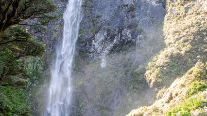 Sunlit waterfall cascades through lush NZ foliage, a serene Arthur's Pass gem