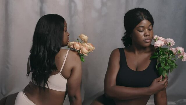 Tilt up shot of two beautiful Black women in underwear sitting on floor in studio and posing for camera with bouquets of roses