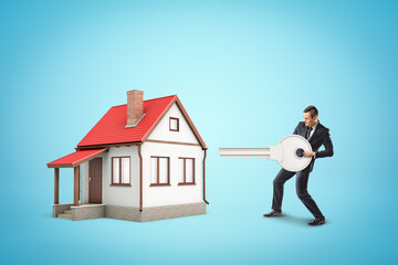 Businessman standing near one-storey cottage and ready to open it with huge key on blue background.
