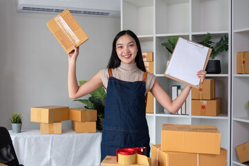 Portrait of young female entrepreneur holding boxes of merchandise and clipboard listing customer...