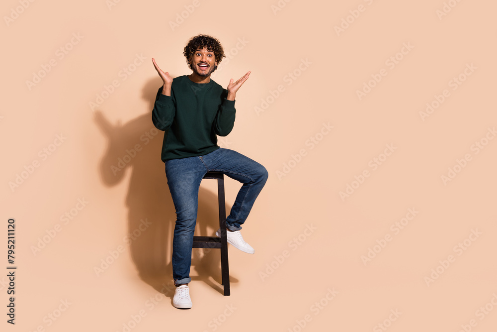 Wall mural Full length photo of positive impressed guy dressed sweatshirt sitting bar chair emtpy space isolated beige color background