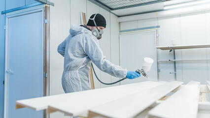 Carpenter with mask using paint spray gun to painting wooden plank in carpentry workshop. High...