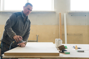 builder carpenter equals polishes wooden board with a random orbit sander in the workshop. High...
