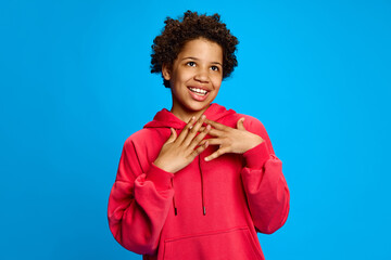Pleasant happy look. Portrait of teen African-American girl in pink hoodie standing with smile, posing against blue studio background. Concept of childhood, human emotions, lifestyle