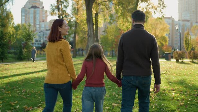 Back view Caucasian happy family weekend holiday at nature outdoors green lawn grass in city autumn park together talking daughter girl child kid go walk parents mother father holding hands walking