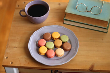 Purple plate filled with pastel macarons, cup of tea or coffee, vintage books and reading glasses on the table. Colorful bookcase in the background. Selective focus.