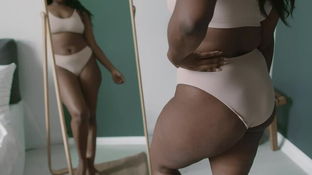 Black woman with curvy body wearing pastel bra and panties looking in mirror and enjoying her beauty uring day at home