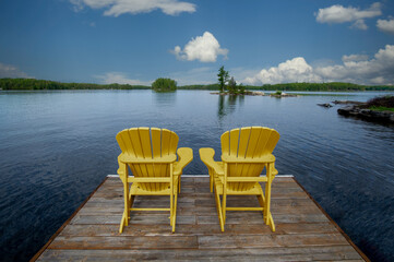 Two wooden Adirondack chairs on a dock greet the tranquil Muskoka morning, overlooking the serene blue lake waters. Across the way, cottages nestled among green trees complete the picturesque scene.
