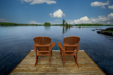 Two yellow Adirondack chairs on a wooden dock in Muskoka, overlooking the tranquil blue lake waters. Across, cottages nestled among green trees complete the scene.