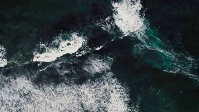 Overhead shot capturing the dynamic motion of white ocean foam against dark rocks as waves break along the coast.