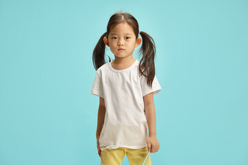 Asian ethnicity little five years old girl, wearing in white t shirt and colorful yellow jeans, has hair is gathered on the sides in a ponytail standing over blue background with a free copy space. - 795251797