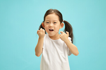 Kid oral hygiene, lovely five years old girl cleaning her teeth using dental floss and looking to camera, having asian ethnciity, wears in white t-shirt, standing against blue isolated. 