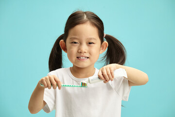 Charming little asian ethnicity child girl squeezes toothpaste out of a tube onto a toothbrush, smiles happily, wears a casual white T-shirt, stands on a blue isolated background. - 795252589