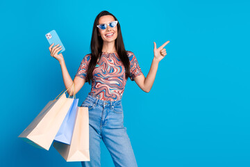Photo of pretty young girl sunglass device shopper point look empty space wear trendy colorful clothes isolated on blue color background