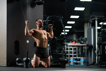 Concentrated handsome sportsman kneeling, while doing exercise for biceps in gym. Low angle view of muscular male athlete in shorts, pulling cables of training apparatus, working out. Sport concept.