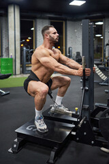 Young man with strong body, squatting with weight, training in modern gym. Side view of handsome Caucasian guy exercising, working out at evening time. Concept of bodybuilding, crossfit, sport.