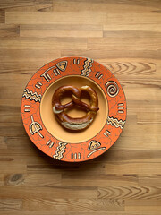 One pretzel lies in one large deep round empty orange plate with ethnic patterns on a wooden background in the center.