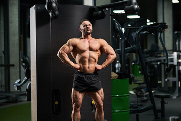 Fototapeta na wymiar Dark-haired bodybuilder performing front lat spread pose in gym. Front view of strong Caucasian male athlete in shorts, lifting chest up, spreading lats and showing to camera. Concept of bodybuilding.