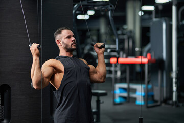 Focused sportsman wearing black tank top, exercising for strong biceps in gym. Portrait of muscular Caucasian bearded guy pulling cables of training apparatus, keeping muscles in tense. Sport concept.