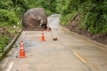Large boulders fall from the mountain, blocking a rural concrete road in a green forest during the rainy season. There are orange cones placed on the road.