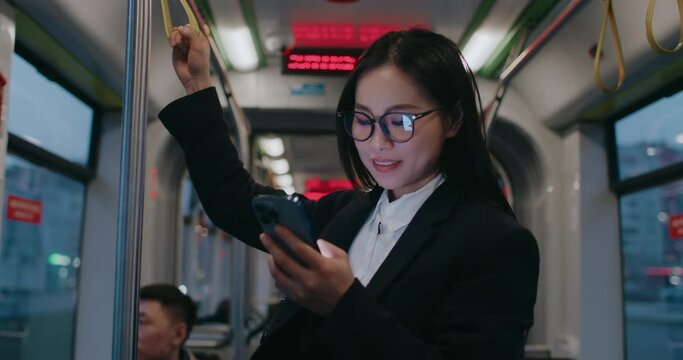 Asian woman holding safety handle while scrolling social media on cell phone on her way back home. Wearing glasses and official clothing. Smiling with joy. Commuting in public transport.