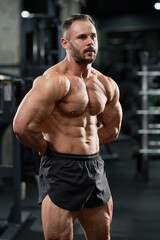 Attractive bearded bodybuilder performing healthy, strong body in gym. Portrait of Caucasian male weightlifter in black shorts, demonstrating muscles, with hands behind back. Concept of bodybuilding.