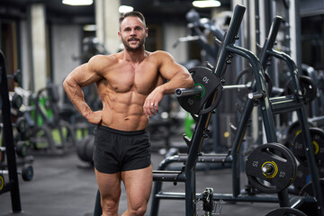 Fototapeta na wymiar Bearded sportsman posing, while leaning on training apparatus in gym. Front view of strong shirtless man smiling to camera, having workout, on blurred background. Concept of bodybuilding, lifestyle.