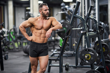 Fototapeta na wymiar Confident shirtless sportsman leaning on training apparatus, having rest after training. Front view of strong guy looking away, while exercising in gym. Concept of bodybuilding, lifestyle, sport.