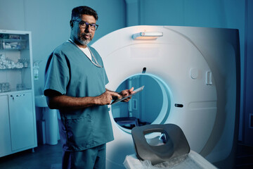 Medium long shot of biracial doctor standing by MRI scanner holding digital tablet looking at...