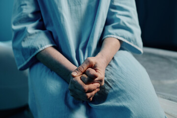 Hands of unrecognizable woman wearing blue patient gown sitting on CT scanner bed in modern clinic,...