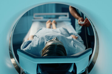 Mature Caucasian woman in blue gown lying on CT scanner bed in modern clinic, unrecognizable doctor...