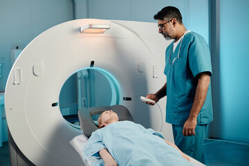 Mature Caucasian woman having CT scan procedure in modern clinic, biracial radiographer holding controller talking to her - Powered by Adobe