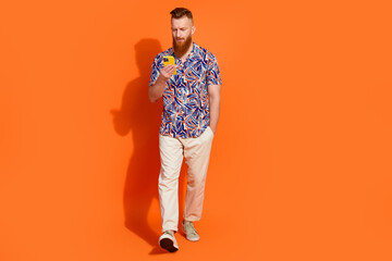 Full length photo of cheerful positive guy dressed print shirt communicating modern device empty space isolated orange color background
