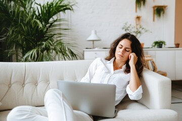 A young female professional looking exhausted while working on a laptop on a cozy home couch. Tired Woman Working Remotely at Home