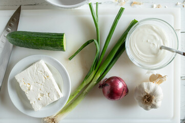Feta cheese, yogurt, onion, garlic, cucumber, spring onion on a cutting board for making a greek...