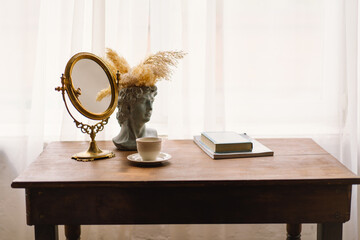 Gold-framed mirror stands on a wooden table, beside a classical bust vase adorned with pampas...