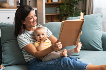 Young happy mother reading a fairytale story book to her baby. Mommy and kid sitting on sofa at home enjoying in imagination. Parent and child lifestyle.