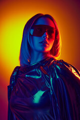 Cyberpunk inspired portrait of young woman in futuristic sunglasses and metallic outfit against gradient orange yellow background in neon. Concept of modern fashion, trends, beauty, youth culture