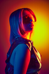Vision of modern elegance. Portrait of young girl in futuristic glasses and metallic dress against gradient orange yellow background in neon light. Concept of modern fashion, trends, beauty, youth