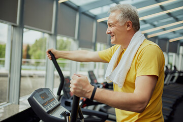 Smiling senior man in vibrant sports attire with towel on shoulders training on exercise machine...