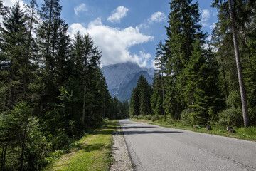 Ampia visuale panoramica sulla strada che attraversa la Val Saisera, circondata da alti alberi verdi, guardando verso le montagne distanti, sotto un cielo parzialmente nuvoloso, di giorno, in estate