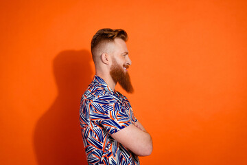 Side profile photo of brutal guy with red beard wear print shirt arms folded look at promo empty space isolated on orange color background