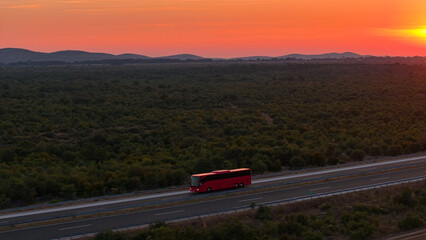AERIAL: A red bus travels down the highway under a captivating crimson sunset. - obrazy, fototapety, plakaty